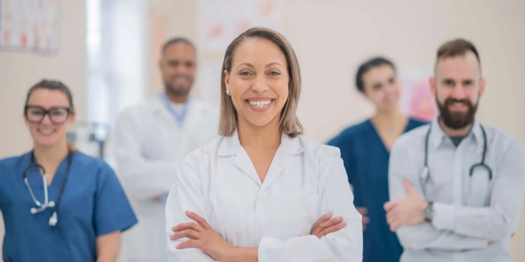 A woman in a white lab coat stands confidently before a group of attentive doctors in a clinical setting.